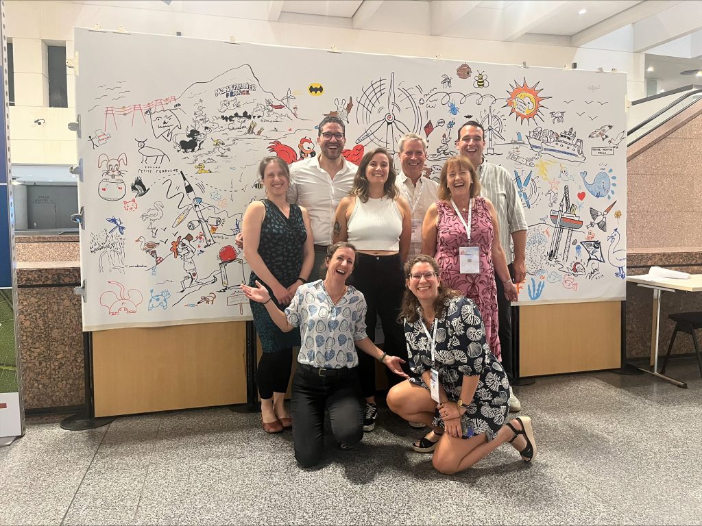 Group photo with the NiD4OCEAN project partners attending CWW25. 8 people attended. Virginie Ramasco (Akvaplan-niva), Juan Pardo (NIVA), Clémentine Mitoyen (CSIC), Arne Myhrvold (EQUINOR), Luca van Duren (Deltares), Pim Somers (North Sea Foundation), Eline van Onselen (North Sea Foundation), Sophie de Reus (North Sea Foundation). They stand in front of the big panel with drawing illustrating the conference and topics discussed.