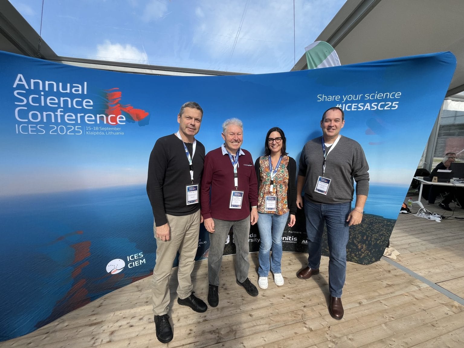 Project partners Marta Moyano, Arturas Razinkovas-Baziukas, David Lusseau, and Darius Daunys, standing in front of a photocall from the Annual Science Conference 2025. The photocall is blue, a photo of the ocean, with the logo of the even on the upper left hand corner